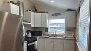 A compact kitchen with a stainless steel refrigerator, a microwave, an oven, and a sink. There is a window with blinds, a decorative sign on the wall, and a small rug on the floor.