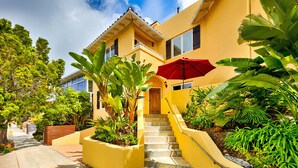Un bâtiment jaune avec un escalier menant à une porte en bois, entouré d’une végétation luxuriante et d’un parasol rouge.