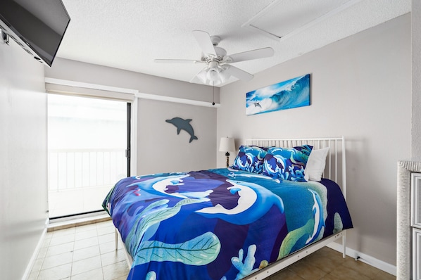 Un dormitorio con cama, ventilador de techo, pintura de delfín y vista al océano a través de la puerta corrediza.