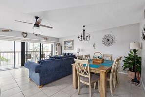 A spacious living area with a blue sofa, wooden dining table, and chairs. There is a ceiling fan, chandelier, and a large window providing natural light. - 4 BR Oasis, water views every room steps to beach! (Clearwater)