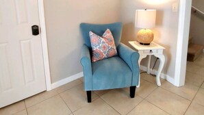 Un sillón azul con cojines estampados, una mesita de noche blanca con una lámpara, y tres cuadros en la pared.