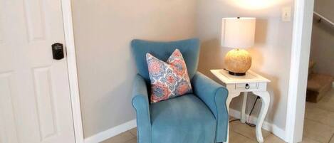 A blue armchair with patterned cushions, a white side table with a lamp, and three framed pictures on the wall.