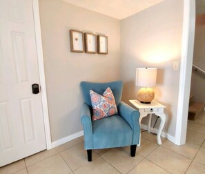 Un sillón azul con cojines estampados, una mesita de noche blanca con una lámpara, y tres cuadros en la pared.