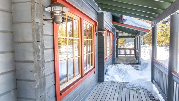 Una terraza de madera con un porche cubierto, un ventanal con detalles rojos y un terreno cubierto de nieve.