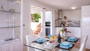 A dining area with a glass table and white chairs, a kitchen in the background, and a view of a garden through an open door. - Villa El Camino by Interhome (Jávea)