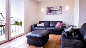 A modern living room with a black leather sofa, an ottoman, and a coffee table. There is a large window with a view of a balcony and greenery. A painting is on the wall, and a floor lamp is on the right side. - Villa El Camino by Interhome (Jávea)