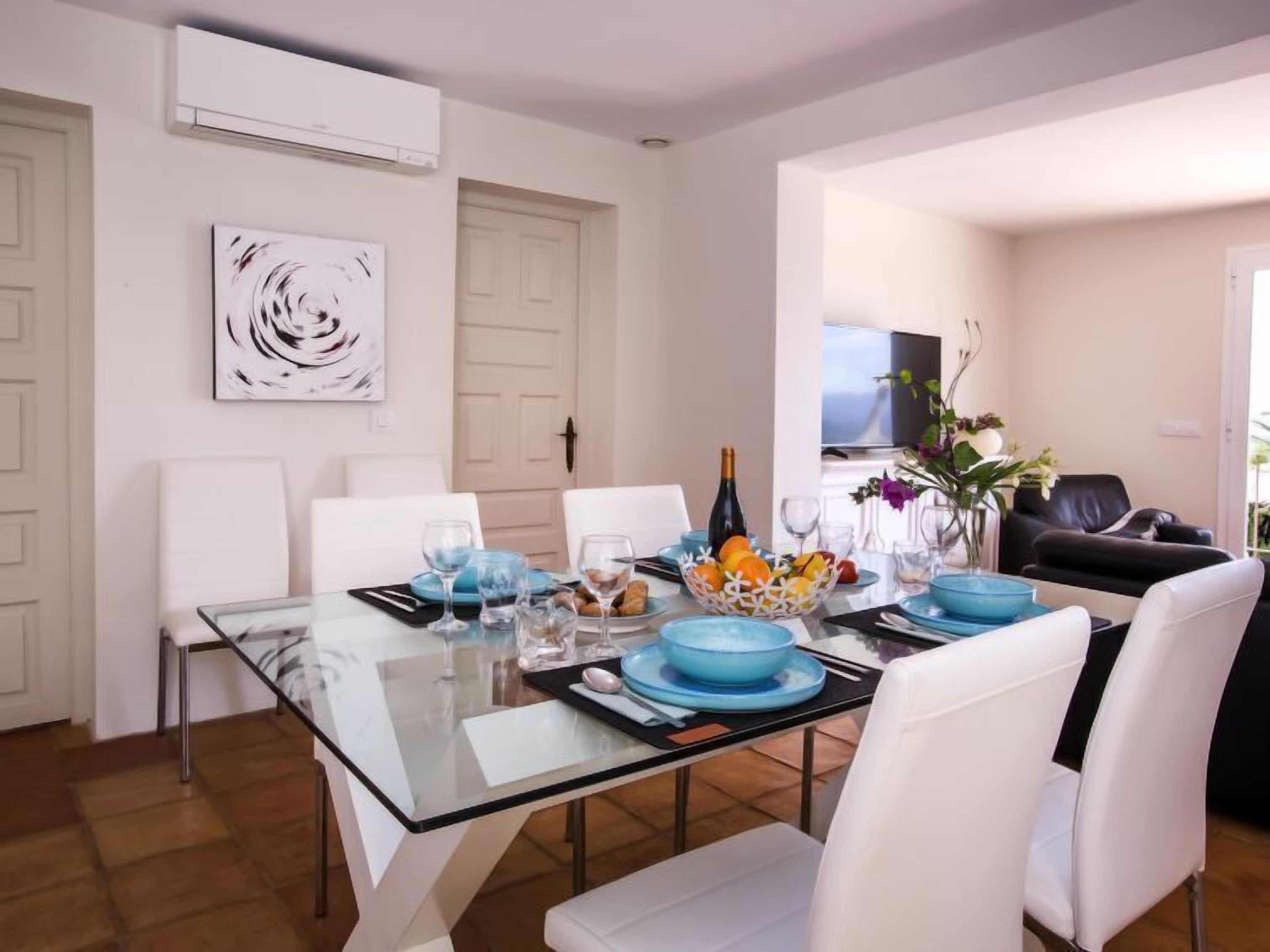 A dining area with a glass table, white chairs, and blue tableware. There is a painting on the wall and a door in the background.