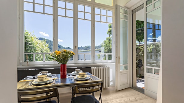 A dining area with a table set for two, a vase with flowers, and a view of greenery and hills through large windows. - Apartment "Garden Vista Lago" in Como with Lake View, Wi-Fi & Private Garden (Como)