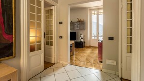 A hallway with a tiled floor, a door with glass panels, a painting on the wall, and a view into a room with a TV and a table. - Vacation Apartment "Central Vista Lago" with Lake View & Private Terrace (Como)