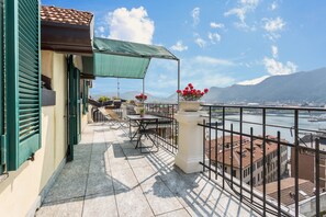 A terrace with a table and chairs, a green awning, and a view of a river and mountains. - Apartment "Attico Vista Lago" in Como with Lake View, Wi-Fi & Terrace (Como)