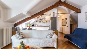 Un salon chaleureux avec un canapé blanc, une table à manger en bois sur laquelle on peut voir des fruits, et une cuisine en arrière-plan.