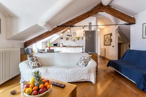Un salon chaleureux avec un canapé blanc, une table à manger en bois sur laquelle on peut voir des fruits, et une cuisine en arrière-plan.