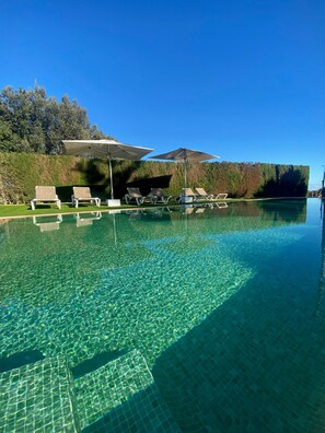 A clear swimming pool with lounge chairs and umbrellas, surrounded by a hedge and trees.