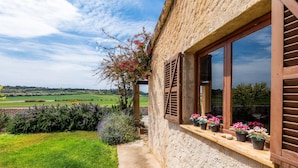 Ein rustikales Steinhaus mit Holzhörnern, ein Garten mit Blumen und Blick auf ein grünes Feld.