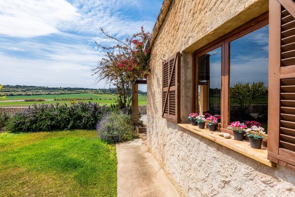 A rustic stone building with wooden shutters, a garden with flowers, and a view of a green field.