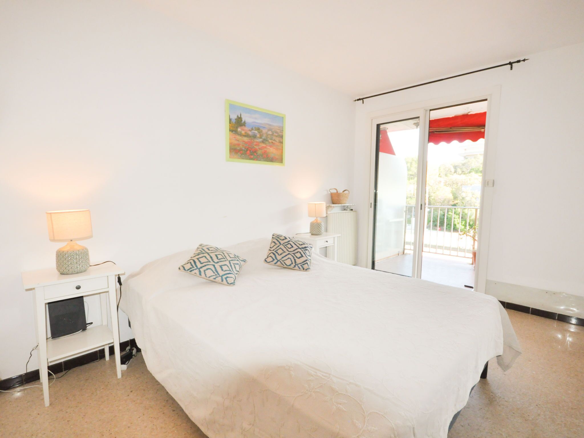 A bedroom with a bed, two bedside lamps, a small white nightstand, a painting on the wall, and a sliding glass door leading to an outdoor area.