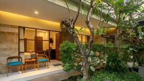 A room with a bed, a desk, and chairs, a small indoor garden with a tree, and a wall-mounted TV. - Naksa Homestay Sanur (Denpasar)