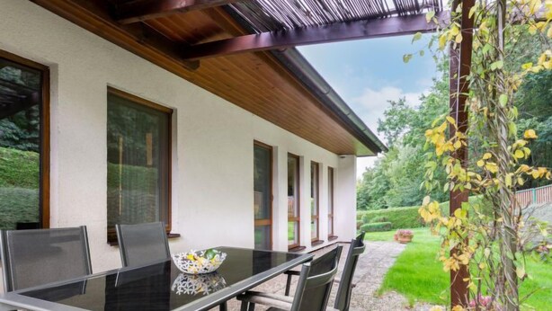 A modern outdoor dining area with a glass table and metal chairs, surrounded by a white wall and large windows. - Vacation home Měřín by Interhome (Slapy)