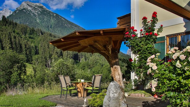 A patio with a wooden table and chairs, surrounded by a garden with flowers and a small pond.