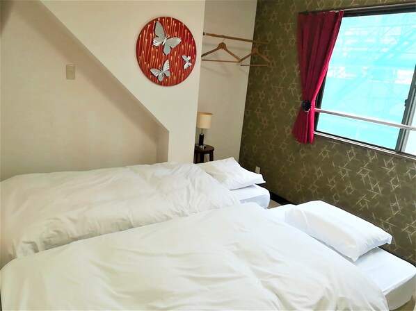 A hotel room with two beds, a wall-mounted circular artwork with butterflies, a window with a red curtain, and a small table with a lamp. - Cote House (Osaka)