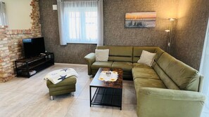 A living room with a green sofa, a small ottoman, a coffee table, a TV on a stand, and a brick wall.