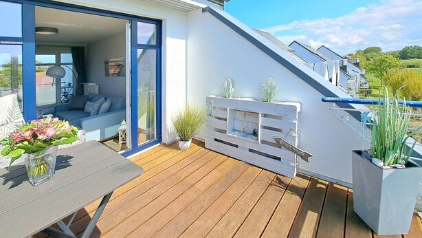 A modern house with a sloped roof, a wooden deck, and a glass door leading to a living area with a sofa and a table. - Haffblick Whg. Ha16, Deutschland (Ostseebad Boltenhagen)