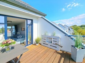 Une maison moderne avec un toit en pente, une terrasse en bois et une porte vitrée donnant sur un espace de vie comprenant un canapé et une table.
