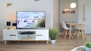 A modern living room with a flat-screen TV, a dining table with chairs, a white cabinet, and a potted plant. - Haffblick Whg. Ha16, Deutschland (Ostseebad Boltenhagen)