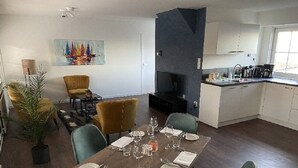 Een eethoek met een houten tafel voor vier personen, omringd door groene stoelen. Aan de muur hangt een schilderij van zeilboten, en er staat een potplant in de hoek.