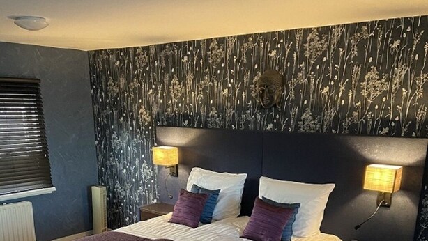 Een groot bed met een donker hoofdbord, twee nachtkastjes met lampen, en een wand met een bloemenmotief.