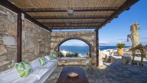 A stone-walled outdoor seating area with a wooden table and cushions, overlooking the sea. - Asimina (Mykonos)