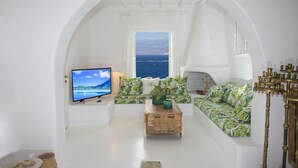 A living room with a television, a sofa, a coffee table, and a view of the sea through an arched doorway. - Asimina (Mykonos)