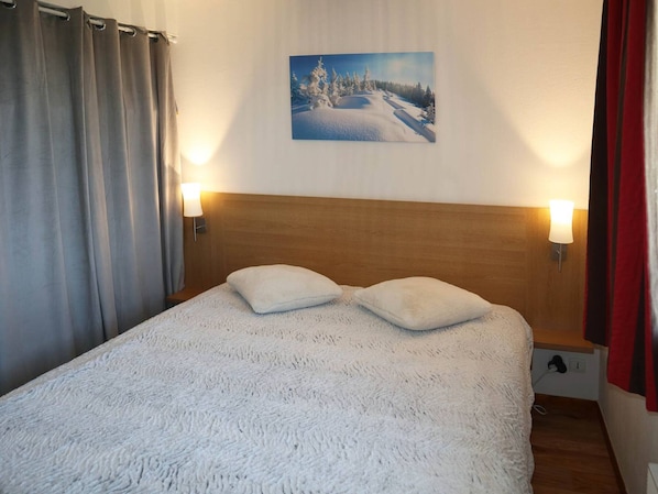 Makuuhuoneessa on suuri sänky, kaksi tyynyä, puinen pääty ja taulu, jossa on lumipeitteinen maisema.