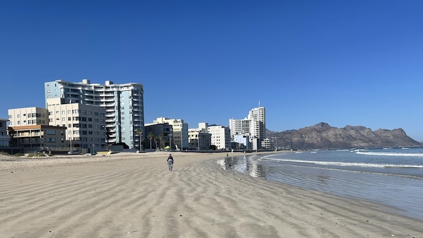 Ein weiter Sandstrand mit einer Person, Gebäuden im Hintergrund und Bergen in der Ferne.
