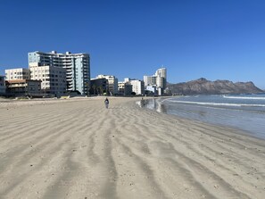 Una extensa playa de arena con una persona caminando, edificios al fondo y montañas a lo lejos.