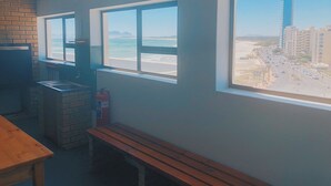 Ein Zimmer mit Backsteinwand, einer Holzbank, einem Waschbecken und einem Fenster mit Blick auf Strand und Gebäude.