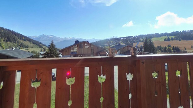 A wooden balcony with a view of a mountainous landscape, including houses and a clear sky.