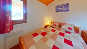 A bedroom with a wooden ceiling, a window with red curtains, a bed with a checkered quilt, and a wall-mounted photo collage.