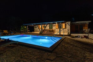 A swimming pool with a stone deck, a covered patio area with outdoor furniture, and a stone building with a satellite dish.