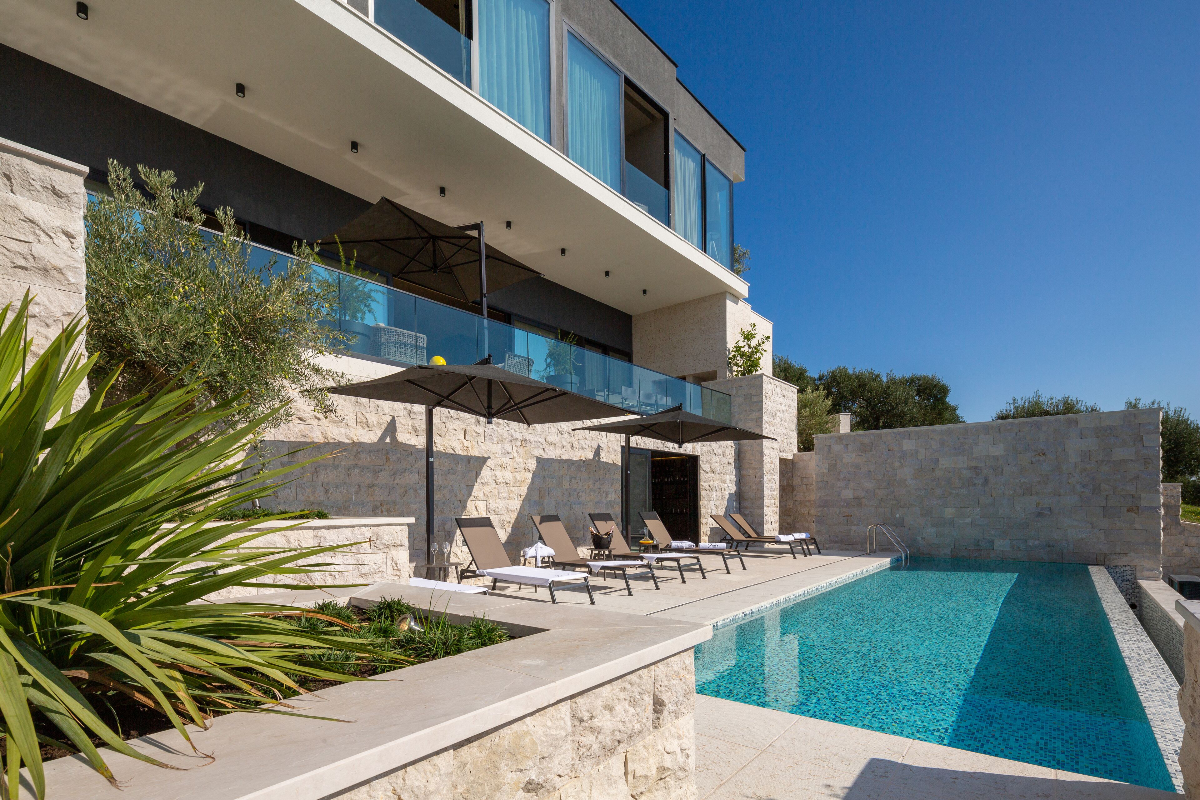 A modern poolside area with lounge chairs and umbrellas, a clear blue pool, and a stone building with a balcony.