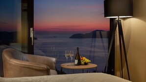 Una habitación con vistas al mar al atardecer, una mesa con una botella y copas, y una lámpara.