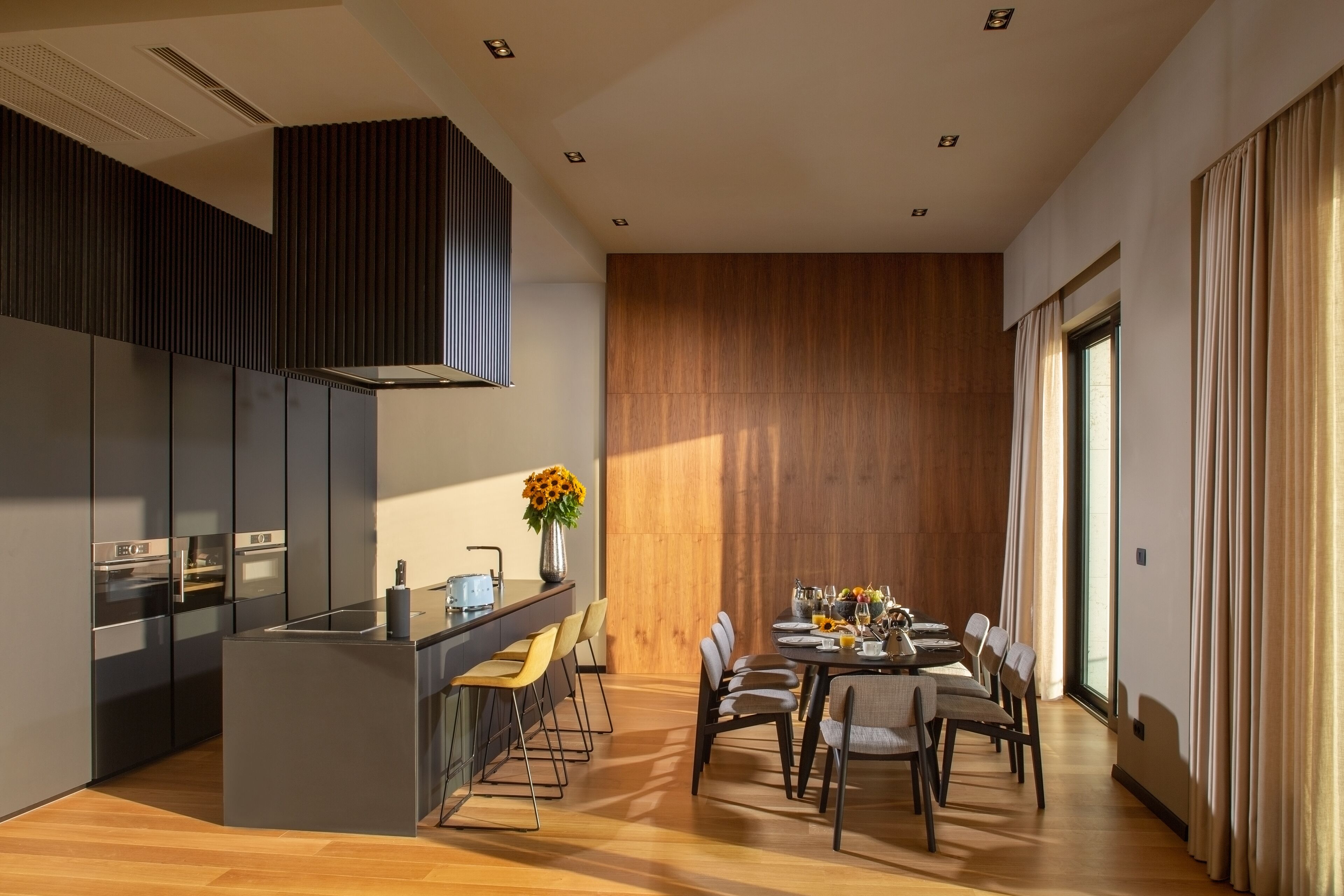 Una cocina moderna con armarios oscuros, una isla central y un comedor con un muro de acento de madera y ventanas amplias.