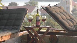 Un balcone con due poltrone reclinabili, un tavolino con una bottiglia di vino e due bicchieri, e vista su un corpo d'acqua ed edifici in lontananza.