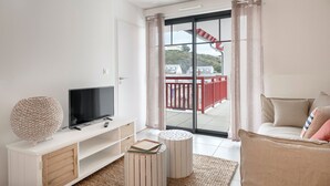 A modern living room with a flat-screen TV, a white entertainment unit, a round ottoman, and a sofa with striped pillows. A large window with a view of a red railing and buildings is visible. - Flat for 4 by the sea in Pléneuf-Val-André (Pléneuf-Val-André)