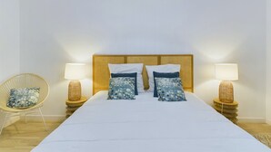 Une chambre à coucher avec un grand lit, deux lampes de chevet, un fauteuil en osier et une tête de lit en fibres tressées.