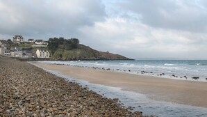 Une plage de galets bordée d’une côte rocheuse, une petite colline avec des bâtiments, et l’océan en arrière-plan.