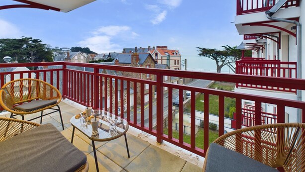 Un balcon agrémenté de fauteuils en osier et d’une table en verre, d’où l’on peut admirer une ville côtière avec ses balustrades et ses bâtiments rouges.