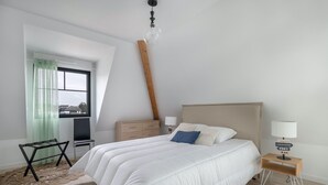 A bedroom with a large bed, a desk, a chair, a lamp, and a view of the city through a window. - Flat for 6 50m from the beach (Pléneuf-Val-André)