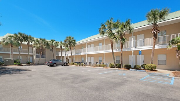 A multi-story motel with a parking lot, palm trees, and a clear blue sky. - ☀️ Close to the Beach- Caribbean Dunes 121 🌴🏖️ (Destin)