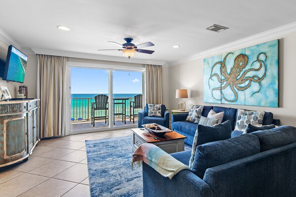 A living room with a view of the ocean, a ceiling fan, and a large octopus painting.
